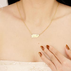 May include: A gold-toned necklace featuring a delicate chain and a small, horizontal rectangular pendant shaped like the island of Puerto Rico. The pendant is engraved with cursive script text and a tiny heart symbol on the right side.