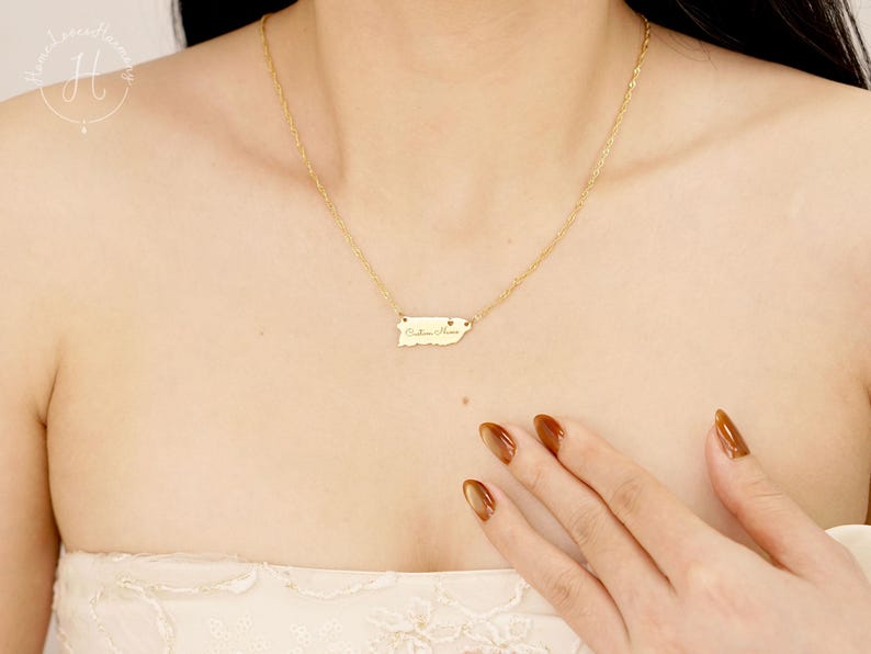May include: A gold-toned necklace featuring a delicate chain and a small, horizontal rectangular pendant shaped like the island of Puerto Rico. The pendant is engraved with cursive script text and a tiny heart symbol on the right side.