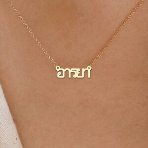 May include: A gold-coloured necklace with a fine chain. The pendant displays a word in a stylized script. The necklace is set against a skin-toned background.