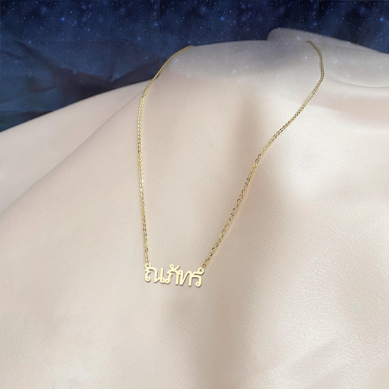 May include: A delicate gold necklace featuring a Thai script pendant. The necklace is presented on a soft, light-coloured fabric, with a dark blue and starry background. The chain is thin and the pendant is in a cursive font.