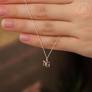 May include: A delicate gold-colored necklace with a pendant featuring the intertwined letters "NG". The necklace is suspended from a fine chain. The background is blurred, highlighting the jewelry.