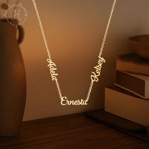 May include: A gold-toned metal necklace featuring three distinct name pendants integrated into the chain. The names Adela, Ernesta, and Kelsey are written in a cursive script font. The chain is a delicate link style, suspending the names.