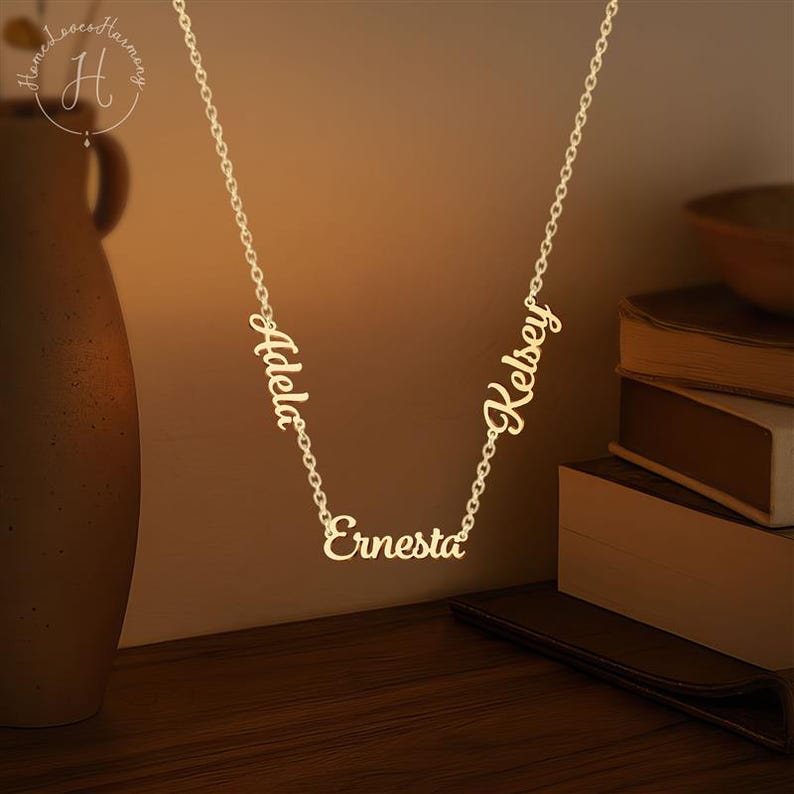 May include: A gold-toned metal necklace featuring three distinct name pendants integrated into the chain. The names Adela, Ernesta, and Kelsey are written in a cursive script font. The chain is a delicate link style, suspending the names.