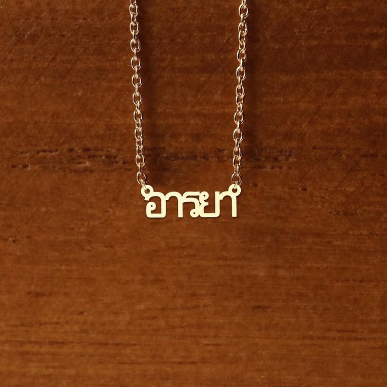 May include: A gold-coloured necklace with a delicate chain. The pendant features the Thai word "อารยา" in a stylized font. The necklace is displayed against a warm, brown wooden background.