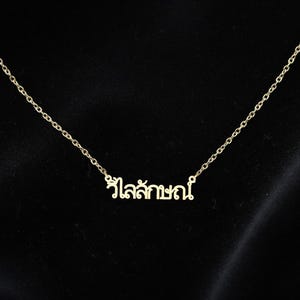 May include: A gold-coloured necklace with a fine chain and a pendant bearing Thai script. The pendant is centrally positioned on the chain. The background is a plain black, which provides a contrast to the necklace.