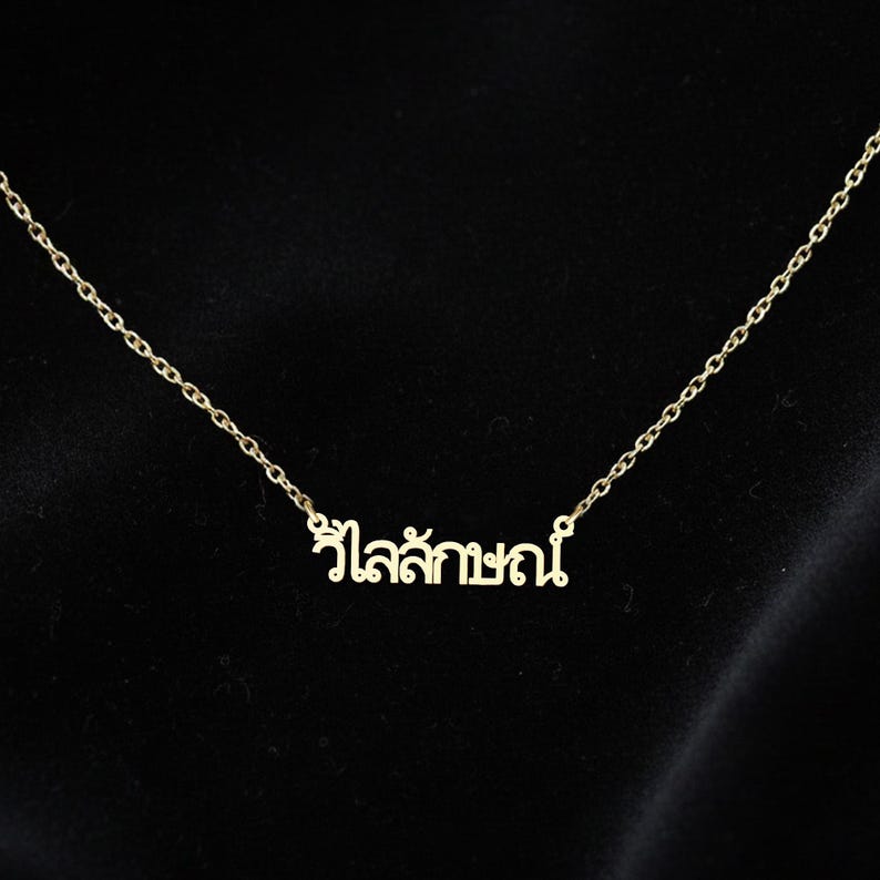 May include: A gold-coloured necklace with a fine chain and a pendant bearing Thai script. The pendant is centrally positioned on the chain. The background is a plain black, which provides a contrast to the necklace.