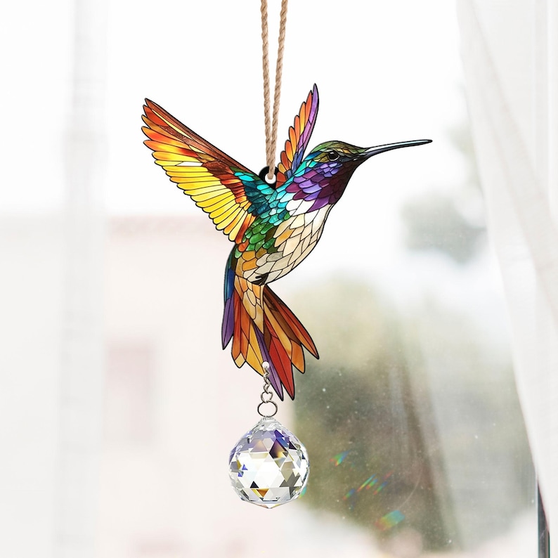 May include: A stained glass hummingbird suncatcher with a crystal prism hanging from it. The hummingbird is colorful with shades of yellow, green, blue, and purple. The crystal prism is clear and reflects light.