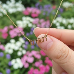 May include: A gold necklace with a fine chain. The pendant displays Thai script. The necklace is held by a hand against a blurred backdrop of pink, white, and purple flowers.