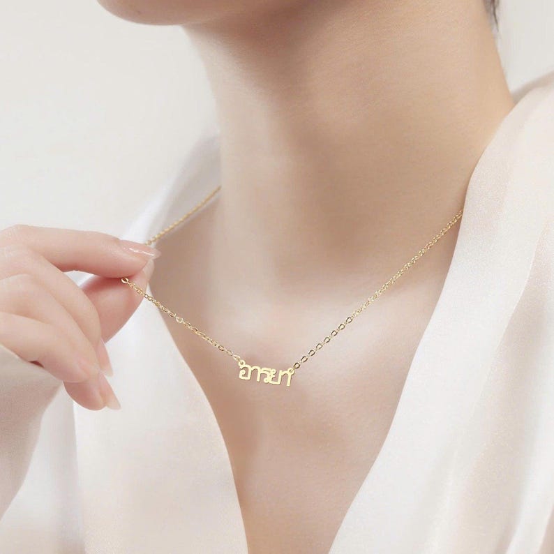May include: A delicate gold necklace with a pendant featuring Thai script. The necklace is worn around the neck. The chain is thin and the pendant is small. The background is a soft, neutral colour.