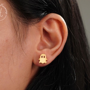 May include: A gold-toned stud earring shaped like a small ghost with two oval-shaped cutouts for eyes and a rounded bottom edge. The earring is positioned on an earlobe, featuring a smooth, polished surface that reflects light across its flat face.