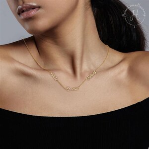 May include: A gold-toned necklace featuring three distinct name pendants integrated into a delicate chain. The names Adela, Ernesta, and Kelsey are written in a cursive script font and are spaced along the length of the thin metal chain.