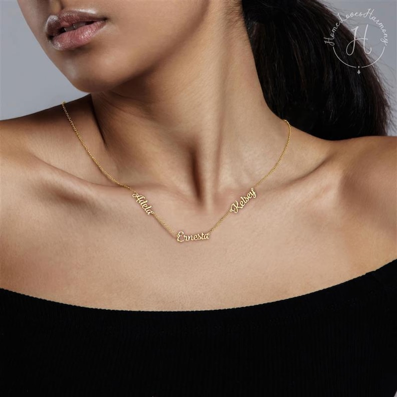 May include: A gold-toned necklace featuring three distinct name pendants integrated into a delicate chain. The names Adela, Ernesta, and Kelsey are written in a cursive script font and are spaced along the length of the thin metal chain.