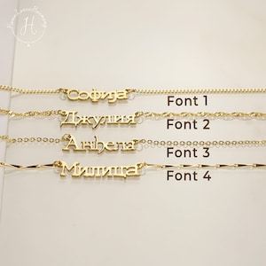Könnte beinhalten: Vier goldfarbene Namensketten, die horizontal angeordnet sind. Jede Kette zeigt einen anderen Schriftstil, beschriftet mit Font 1, Font 2, Font 3 und Font 4. Die Namen sind in kyrillischen und lateinischen Buchstaben an zarten goldfarbenen Metallketten.