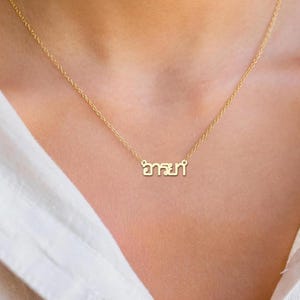 May include: A gold-coloured necklace with a fine chain. The pendant displays a word in a stylized script. The necklace is worn against a person's skin and a white shirt.