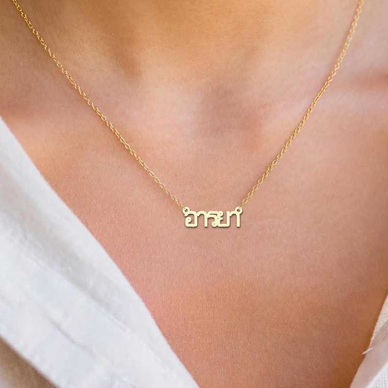 May include: A gold-coloured necklace with a fine chain. The pendant displays a word in a stylized script. The necklace is worn against a person's skin and a white shirt.