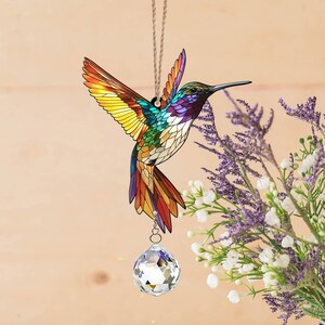 May include: A stained glass hummingbird suncatcher with a crystal prism hanging from its beak. The hummingbird is colorful with shades of yellow, green, blue, and red. The suncatcher is hanging from a string.
