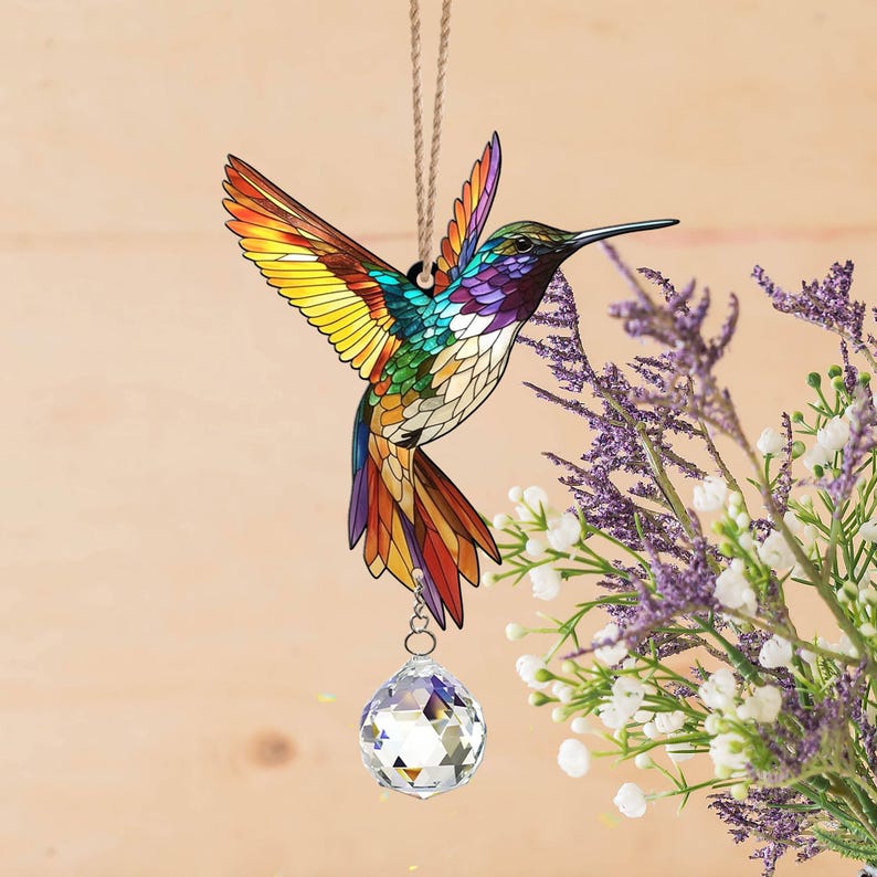 May include: A stained glass hummingbird suncatcher with a crystal prism hanging from its beak. The hummingbird is colorful with shades of yellow, green, blue, and red. The suncatcher is hanging from a string.