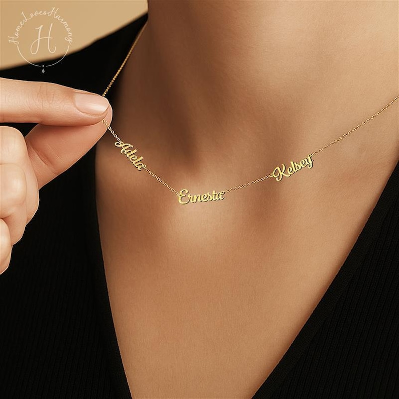 May include: A gold-toned chain necklace featuring three distinct name pendants in a cursive script font. The names Adela, Ernesta, and Kelsey are integrated directly into the delicate metal chain, creating a personalized jewelry piece.