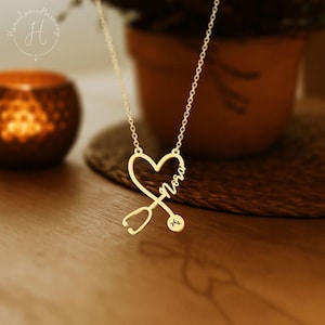 May include: A gold-colored necklace with a heart-shaped pendant. The pendant features a stethoscope design, the name "Nora" in cursive, and a small heart rate symbol. The necklace is on a wooden surface.