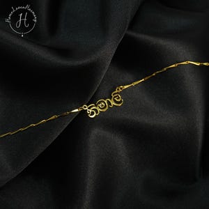 May include: A delicate gold-coloured necklace with a name pendant in a cursive font. The necklace is displayed on a black satin fabric. The brand logo "Home Loves Harmony" is in the upper left corner.