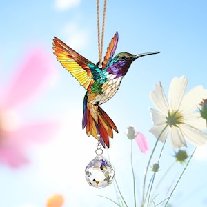 May include: A stained glass hummingbird suncatcher with a crystal prism hanging from it. The hummingbird is colorful with shades of red, orange, yellow, green, blue, and purple. The suncatcher is hanging from a string against a blue sky background.