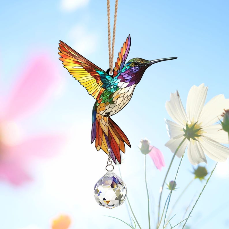 May include: A stained glass hummingbird suncatcher with a crystal prism hanging from it. The hummingbird is colorful with shades of red, orange, yellow, green, blue, and purple. The suncatcher is hanging from a string against a blue sky background.