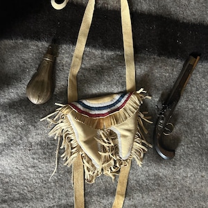 Puede incluir: Una bolsa de cuero marrón con flecos y una parte superior con cuentas, un frasco de pólvora plateado y una réplica de pistola de chispa se exhiben sobre una manta gris. La bolsa tiene una correa larga y está decorada con cuentas rojas, blancas y azules.