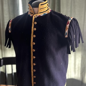 Op de afbeelding: Een zwarte wollen vest met gouden randen en knopen. Het vest heeft een hoge kraag met gouden strepen en kleurrijk kralenwerk. De schouders hebben decoratief kralenwerk en zwarte franjes.