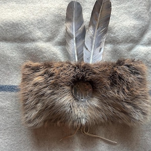 Könnte beinhalten: Ein braunes Fellstirnband mit zwei grauen und weißen Federn. Das Stirnband hat einen kreisförmigen silbernen Knopf mit einem geätzten Design und einen Lederriemen. Es wird auf einer grau-blau gestreiften Decke präsentiert.