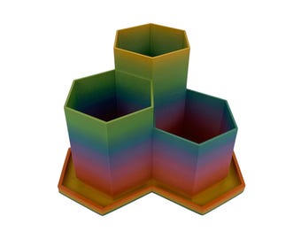 May include: A desk tidy featuring three hexagonal compartments of varying heights arranged on a matching hexagonal base. The object displays a vibrant gradient colour transition shifting from yellow at the top to orange, red, purple, blue, and green.