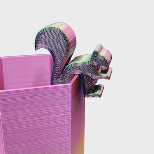 May include: A pink rectangular container with a multi-colored squirrel-shaped accent. The squirrel is perched on the edge of the container, with a textured surface and a gradient of colors including pink, green, and blue. The container has a layered appearance.
