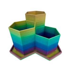May include: A multi-compartment desk organizer featuring three hexagonal containers of varying heights arranged on a matching hexagonal base. The object displays a gradient color pattern transitioning from yellow at the top to green, blue, and purple at the base.
