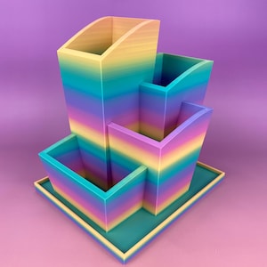 Könnte beinhalten: Ein Mehrfach-Schreibtisch-Organizer in einem Regenbogen-Farbverlauf. Der Organizer hat drei unterschiedlich hohe und große Abschnitte, alle mit rechteckiger Basis. Die Farbe geht von Gelb über Blau zu Violett über.