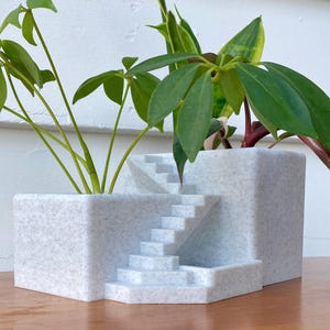 May include: A unique, speckled gray planter with a built-in staircase design. The planter holds several green plants with various leaf shapes. The planter sits on a wooden surface, with a white wall in the background.