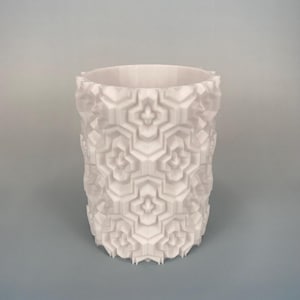 May include: A white cylindrical vase with a geometric pattern. The vase has a textured surface with a repeating design of stylized floral shapes. The vase is made of a solid white material and stands on a light blue background.