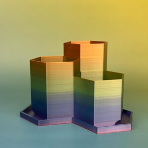 May include: A multi-tiered, geometric desk organizer with a gradient color scheme. The organizer features three hexagonal compartments of varying heights, transitioning from blue to green, yellow, and orange. The base is a matching color with a red trim.