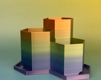 May include: A multi-tiered, geometric desk organiser with a gradient colour scheme. The organiser features three hexagonal compartments of varying heights, transitioning from blue to green, yellow, and orange. The base is a matching colour with a red trim.