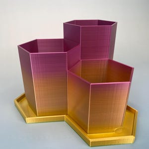 Könnte beinhalten: Ein Set aus drei sechseckigen Pflanzgefäßen in einem Farbverlauf, der von einem tiefen Magenta oben zu einem goldgelben Farbton an der Basis übergeht. Die Pflanzgefäße stehen auf einem passenden sechseckigen Tablett.