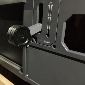 May include: Close-up of a dark gray control panel with a black knob and a lever. The panel has the words "UP" and "DOWN" with directional arrows. The knob is textured, and the lever is in the middle position.