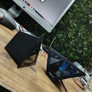 Può includere: Due componenti elettronici neri di forma geometrica con superfici chiare e riflettenti e cablaggio visibile sono esposti su una superficie di legno. Uno è un prisma rettangolare, l'altro un prisma triangolare.