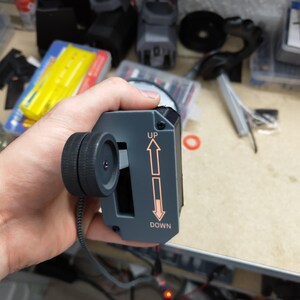 May include: A gray electronic device with a black wheel and the words "UP" and "DOWN" with arrows. The device has a black cord and is held in a hand. The background is a cluttered workbench.