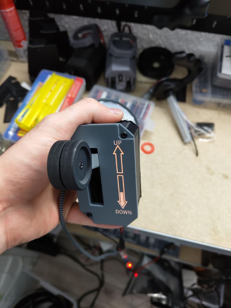 May include: A gray electronic device with a black wheel and the words "UP" and "DOWN" with arrows. The device has a black cord and is held in a hand. The background is a cluttered workbench.