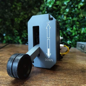 May include: A grey control lever with a black wheel and the words "UP" and "DOWN" with arrows. The lever is set against a backdrop of green foliage and a wooden surface.
