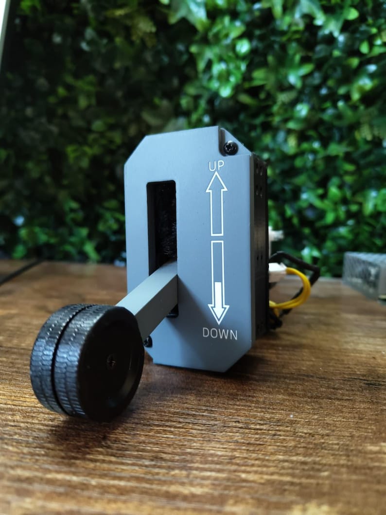 May include: A gray control lever with a black wheel and the words "UP" and "DOWN" with arrows. The lever is set against a backdrop of green foliage and a wooden surface.