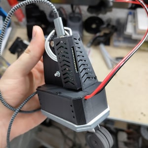 May include: A black electronic device with a gray and white braided cable and a red and black wire. The device has a rectangular base with a small wheel and a slotted top section. The device is held in a person's hand.
