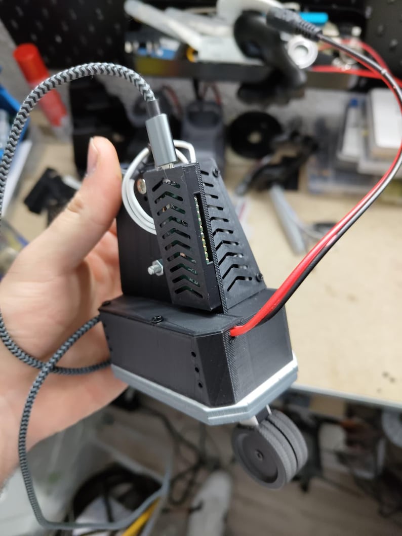 May include: A black electronic device with a gray and white braided cable and a red and black wire. The device has a rectangular base with a small wheel and a slotted top section. The device is held in a person's hand.