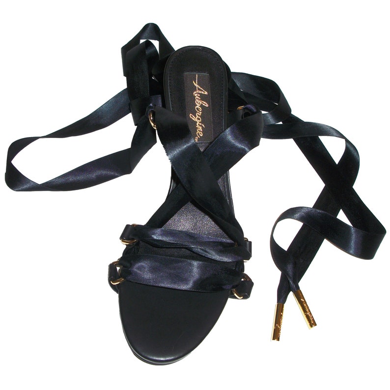 Op de afbeelding: Zwarte satijnen sandalen met vetersluiting en gouden accenten, met de merknaam "Aubagio" op de binnenzool.
