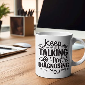 May include: White ceramic coffee mug with black text that reads "Keep Talking I'm Diagnosing You". The mug is sitting on a wooden table with a laptop and a plant in the background.
