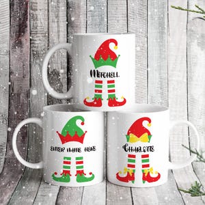 Può includere: Tre tazze in ceramica bianca con disegni di elfi di Natale. Le tazze hanno gambe e cappelli di elfo rossi e verdi. Le tazze sono personalizzate con i nomi "Mitchell", "Inserisci il nome qui" e "Charlotte".