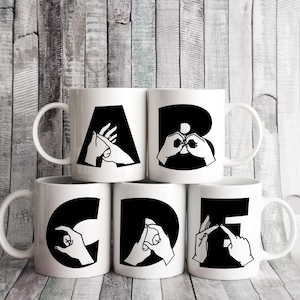 Könnte beinhalten: Fünf weiße Keramikbecher mit schwarzen Griffen. Jeder Becher zeigt einen anderen Buchstaben des Alphabets, A, B, C, D und E, dargestellt durch ein Zeichen der Gebärdensprache.